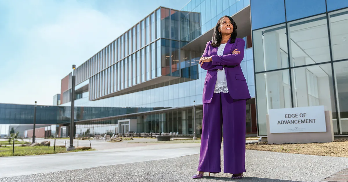 President Angela L. Walker Franklin, PhD, stands outside DMU’s Edge of Advancement building with arms crossed, wearing a purple suit. The modern glass-fronted building and campus walkways are visible behind her, with an "Edge of Advancement" sign in the foreground.