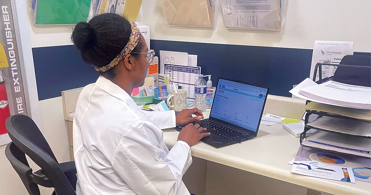 Tiphani James, DPM’16, sits at a workstation in a clinical setting, using a laptop at a desk with patient care materials and office supplies nearby.