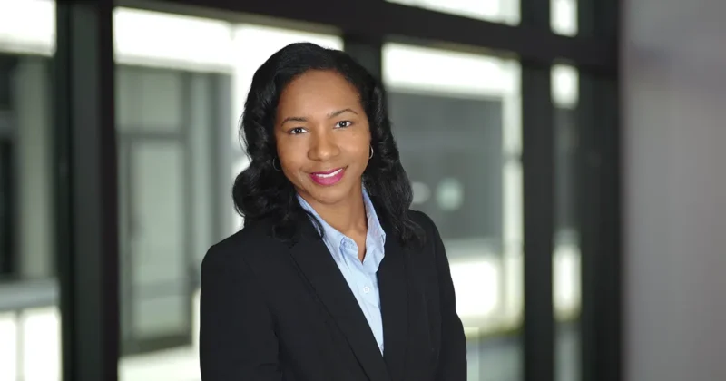 Professional portrait of Tiphani James, DPM’16, wearing a dark blazer and light blue shirt, standing in front of a softly blurred indoor background.