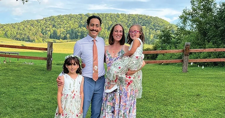 Caitlin Suginaka, MPH’13, stands with her husband, Alex Suginaka, DO’14, and their two children on a grassy field in front of a wooden fence and tree-covered hills. The family poses outdoors beneath a blue sky with scattered clouds.