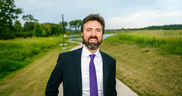 Jason Semperini stands on a paved path beside a grassy landscape and waterway, wearing a dark suit and purple tie.